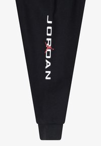 Fekete tréningnadrág puha anyagból, fehér "JORDAN" felirattal és egy piros áthúzott logóval az oldalon, a bokánál pedig szűkített mandzsetták találhatók.