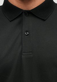 Black polo shirt with a textured collar, two button placket, and a pocket. Fabric has a smooth finish with a subtle weave pattern.