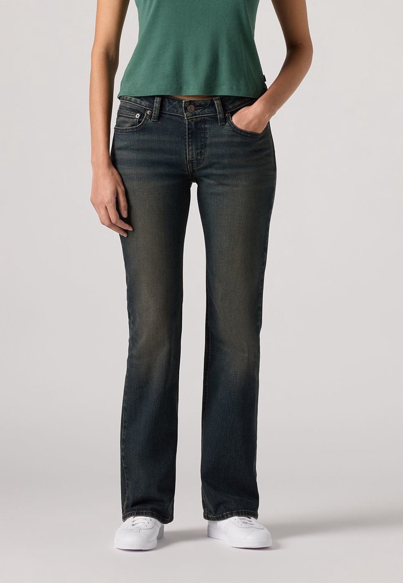 Personne portant un jean bootcut foncé délavé, un crop top vert et des baskets blanches, debout avec une main dans la poche.