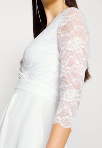 Weißes Kleid mit langen Spitzenärmeln, das einen figurbetonten Oberteil und einen fließenden Rock hat. Die Spitze weist florale Muster und eine strukturierte Optik auf.