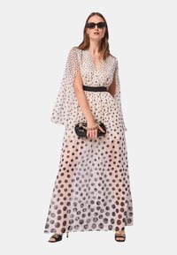 Abito maxi a pois in tessuto trasparente con vita segnato da una cintura, caratterizzato da grandi pois neri su uno sfondo chiaro e maniche a sbuffo. Clutch nera e sandali.