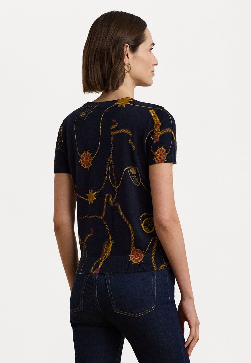 Niebieski sweter z krótkim rękawem z złotymi i czerwonymi wzorami w marynarskim stylu. Posiada prążkowany kołnierz i dół oraz gładką teksturę materiału.