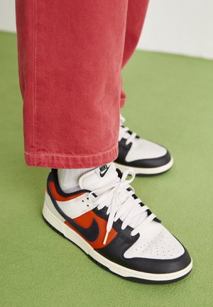 Tenisky Nike Dunk mají bílé kožené podšívky s černými a oranžovými akcenty, kulatou špičku a bílé tkaničky. Jsou kombinovány s červenými kordurovými kalhotami.