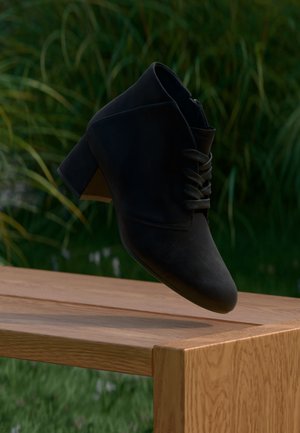 Bottine noire en daim avec talon carré et lacets flottant au-dessus d’un banc en bois à l’extérieur avec des plantes vertes en arrière-plan.