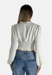 Weißes, kurzes Blusenoberteil mit langen Puffärmeln und gerafftem Rückendetail, kombiniert mit hoch taillierten blauen Jeans aus Denim. Sanfte Textur.