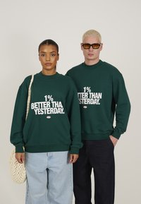 Grønne bomullssweatshirts med "1% BEDRE ENN I GÅR" trykket i hvitt. Modell som har på seg lyseblå jeans og bærer en vevd bag.