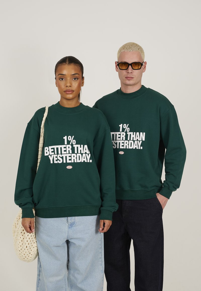 Grønne bomullssweatshirts med "1% BEDRE ENN I GÅR" trykket i hvitt. Modell som har på seg lyseblå jeans og bærer en vevd bag.