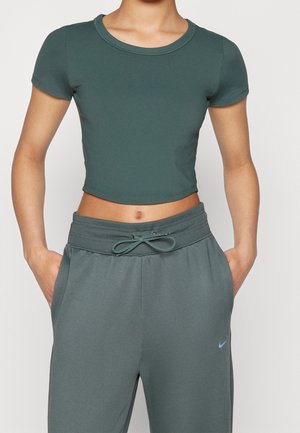 Persoon draagt een donkergroene cropped korte mouw shirt en bijpassende groene joggingbroek met handen in de zakken, toont de buik en een casual stijl.