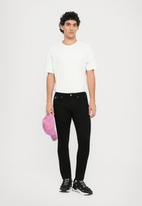 Melni šauri džinsi, baltā T-kreklā un melnās kedās. Tur rokās rozā cepure ar tekstu, džinsiem ir zelta pogas. Rūpīgi teksturēta auduma virsma.