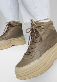 Baskets montantes en cuir marron avec semelles épaisses beige, œillets dorés et lacets, portées avec un jean bleu clair à ourlet effiloché.