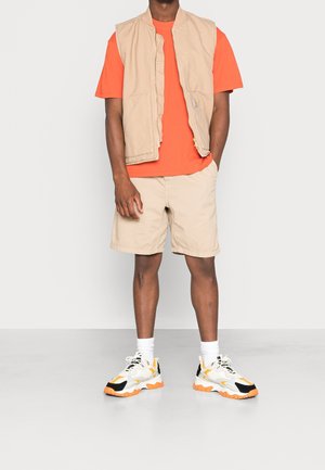 Gilet sans manches beige, t-shirt orange, shorts beiges et baskets noir-orange. Texture de tissu uni, coupe décontractée, design minimaliste.