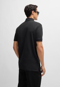 Black cotton polo shirt with short sleeves, a collar, and side slits. Smooth texture and tailored fit, shown from the back. Sunglasses worn.