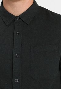 Dunkelgrünes Hemd aus strukturiertem Stoff, mit Kragen, durchgehender Knopfleiste und einer einzelnen Brusttasche.