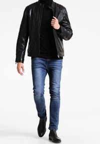 Svart lädermotorcykeljacka med kviltade axeldetaljer, bärs över en svart polotröja, kombinerad med blå slim-fit jeans och svarta ankelboots.