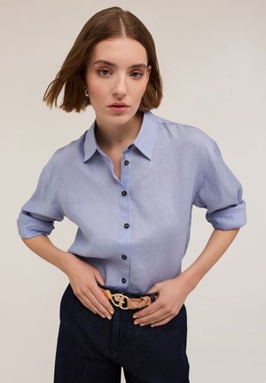 Lichtblauw linnen shirt met een knoopsluiting aan de voorkant, korte opgerolde mouwen en zwarte knopen, gedragen met donkere denim en een riem met gouden hardware.