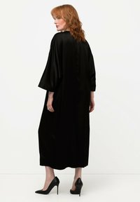 Abito lungo in satin nero con maniche morbide, scollatura a V e silhouette dritta. Indossato con scarpe con tacco alto nere. Texture liscia, accenti minimi.
