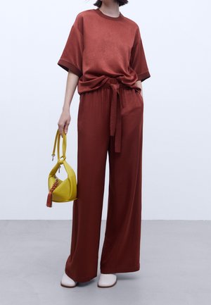 Roodbruine oversized shirt met korte mouwen, geknoopt in de taille, gecombineerd met een broek met wijde pijpen. Houdt een gele handtas met een franjeretail.