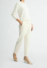 Bílý pletený svetr s akcenty na ramenou, doplněný krémovými zúženými kalhotami s kapsami a zlatými knoflíky, a zlatými podpatky.