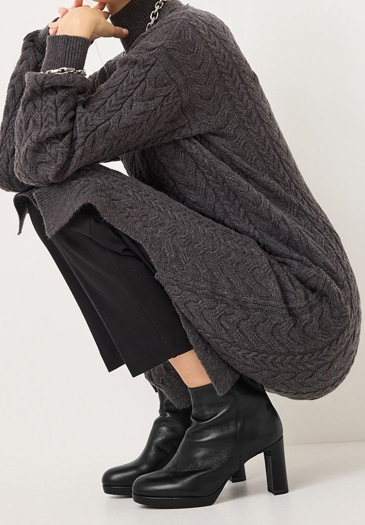 Personne accroupie portant une robe pull torsadée gris foncé, un pantalon court noir, des bottines noires à talons et des bijoux argentés en chaîne.