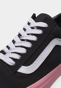 Baskets noires avec lacets blancs, bande latérale blanche et semelle rose, présentées en gros plan sur un fond clair.