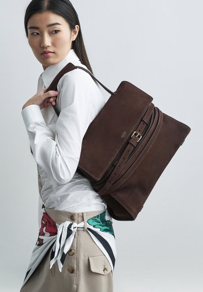 Junge Frau in einem weißen Hemd mit beigem Rock und einem bunten Schal, der um die Taille gebunden ist, trägt einen großen, dunkelbraunen Lederrucksack.