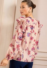 Gėlių blusa iš šviesiai rožinės medžiagos, turinti raukšlėtus pečius, ilgas rankoves ir ryškius mėlynus, raudonus bei žalius gėlių raštus.