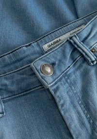 Ανοιχτό μπλε τζιν ύφασμα με κοντινό πλάνο της μέσης, μεταλλικό κουμπί και λευκή ετικέτα που γράφει "GABBA" σε τζιν παντελόνια.