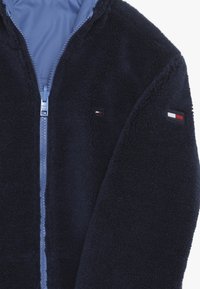 Granatowa kurtka z polaru na zamek, o miękkiej fakturze. Posiada jasnoniebieską podszewkę oraz małe naszywki z logo w kolorach czerwonym, białym i niebieskim na rękawie.