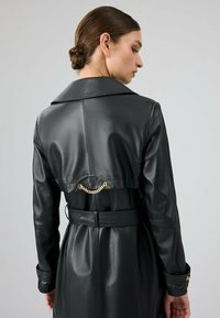 Zwarte leren trenchcoat met een brede kraag, een ceintuur in de taille en een gouden kettingaccent op de rug voor een extra detail.