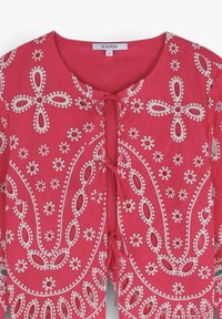 Cardigan rose vif présentant une broderie florale blanche, des découpes rondes et ovales, et une fermeture à nouer au niveau du col. Le tissu semble léger.