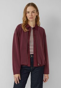 Eine Frau mit hellbraunem Haar trägt eine kastanienbraune, geknöpfte Jacke über einem gestreiften Shirt und dunklen Jeans, während sie vor einem schlichten grauen Hintergrund steht.