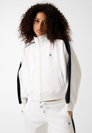 Giovane donna con capelli ricci che indossa una tuta bianca con strisce nere, con le mani nelle tasche, che guarda verso la fotocamera su sfondo bianco.