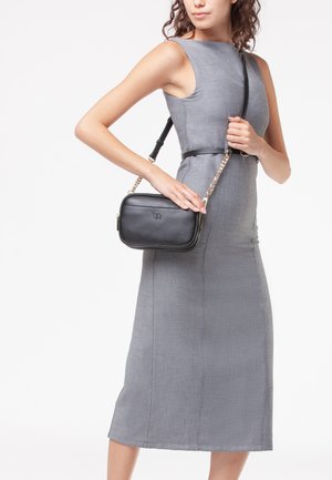 Borsa a spalla nera in pelle con tracolla a catena, forma rettangolare, superficie texturizzata e dettaglio del logo sulla parte anteriore. Indossata con un vestito grigio.