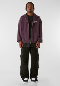 Sudadera morada con cremallera, con bolsillos frontales y un logotipo bordado, combinada con pantalones cargo negros con bolsillos de parche y zapatillas blancas.