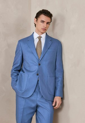 Young man with curly hair wearing a blue suit, white shirt, and brown patterned tie, standing against a beige textured wall.
