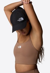Top corto ajustado marrón con el logo de The North Face y una gorra negra con el mismo logo, llevada por una persona con el pelo largo recogido.