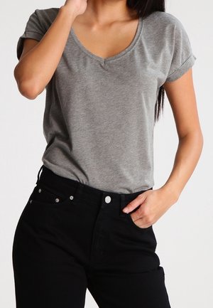 Kvinde iført en grå kortærmet V-hals T-shirt indstukket i sorte jeans med højtaljet, med den ene hånd i lommen og den anden nær ansigtet.