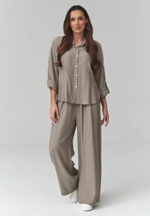 Kobieta z długimi falującymi włosami, ubrana w luźną, taupe'ową koszulę na guziki i pasujące szerokie spodnie, stojąca na tle jednolitego szarego tła.