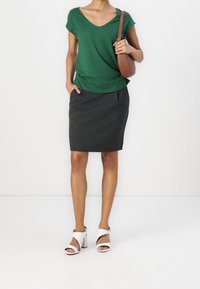 Zelený bavlněný top s výstřihem do V v kombinaci s tmavě šedou tužkovou sukní. Modelka má na sobě bílé sandály na podpatku a drží hnědou crossbody kabelku.