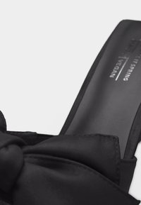 Μαύρο παπούτσι μύλος με μεγάλο υφασμάτινο φιόγκο στην κορυφή και κείμενο ορατό μέσα στην περιοχή της φτέρνας σε ανοιχτό φόντο.