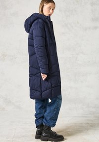 Námořnická péřovka s kapucí, dlouhým střihem, prošívaným designem, zapínáním na zip a postranními kapsami, spojená s modrými džíny a černými botami.