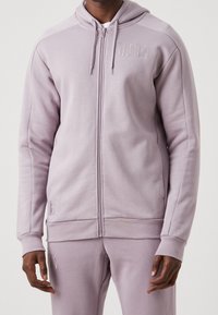 Sudadera con capucha y cremallera de color lavanda claro, con capucha ajustable mediante cordón, textura suave, puños acanalados y un pequeño logo en el pecho izquierdo.