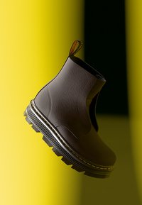 Bottines en cuir marron avec une surface texturée, des panneaux latéraux élastiques et une semelle noire épaisse avec une ligne accentuée jaune.