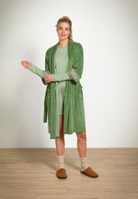 Grüner Velourbademantel mit floral bedruckten Manschetten, zwei Fronttaschen, über einem hellgrünen Kleid getragen. Das Model trägt beigefarbene Hausschuhe und beige Socken.
