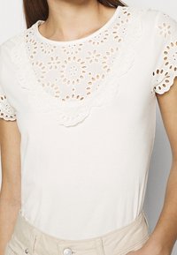 Biała bluzka z krótkim rękawem o ozdobnym, wyciętym dekolcie z haftem typu ażurowego i wzorami kwiatowymi. Miękki materiał o dopasowanym kroju.