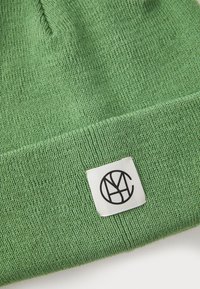 Groene gebreide beanie van zacht materiaal, met een omgeslagen rand en een geweven label met een zwart logo op een witte achtergrond.