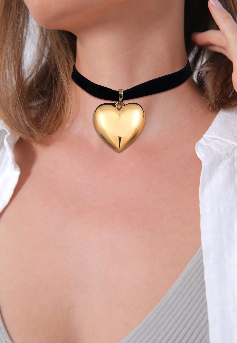 Gros plan d'une personne portant un collier ras du cou en velours noir avec un pendentif brillant en forme de cœur doré au cou.