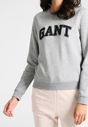 Femme portant un sweat-shirt gris clair avec le logo GANT en lettres noires, associé à un pantalon beige clair, sur un fond uni.