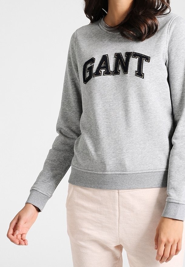 Femme portant un sweat-shirt gris clair avec le logo GANT en lettres noires, associé à un pantalon beige clair, sur un fond uni.