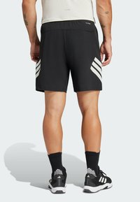 Schwarze Sportshorts mit weißen Seitenstreifen, aus atmungsaktivem Material und elastischem Bund. Getragen mit schwarzen Socken und Schuhen.